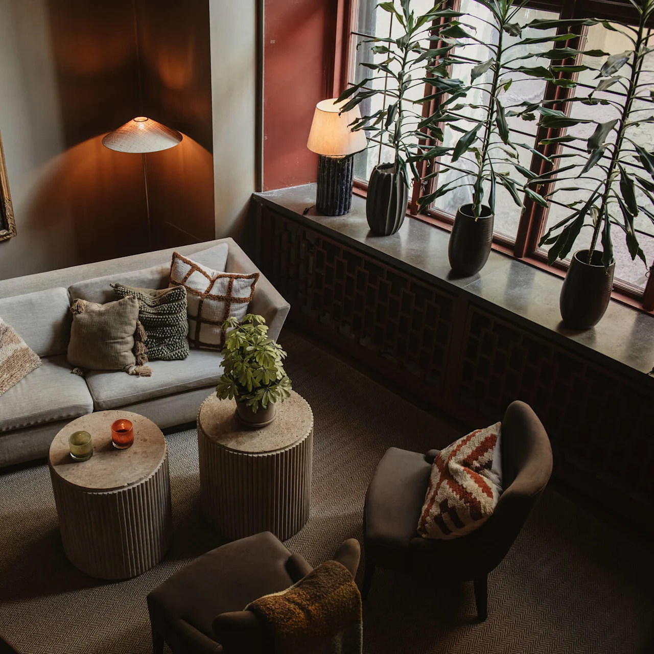 A cozy living room with a beige sofa adorned with cushions, two round tables, a floor lamp, tall potted plants by a window, and artwork on the wall.