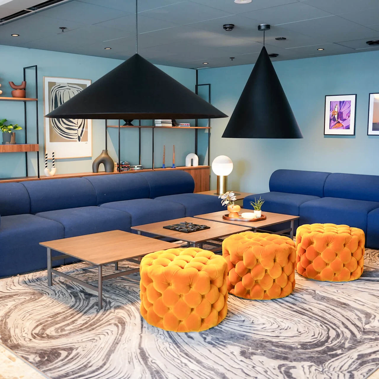 Blue sofas sit on a swirling-patterned rug, while orange, tufted stools are positioned around wooden tables; black cone-shaped lights hang above in a modern lounge space.