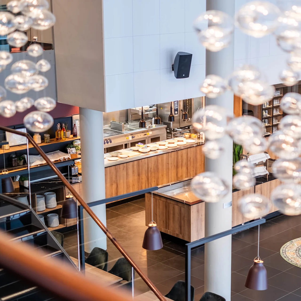 Modern dining area with wooden furniture and decorative glass-like bubbles hanging from the ceiling; an open kitchen visible with plates and utensils; plants and large wall art enhance the atmosphere.