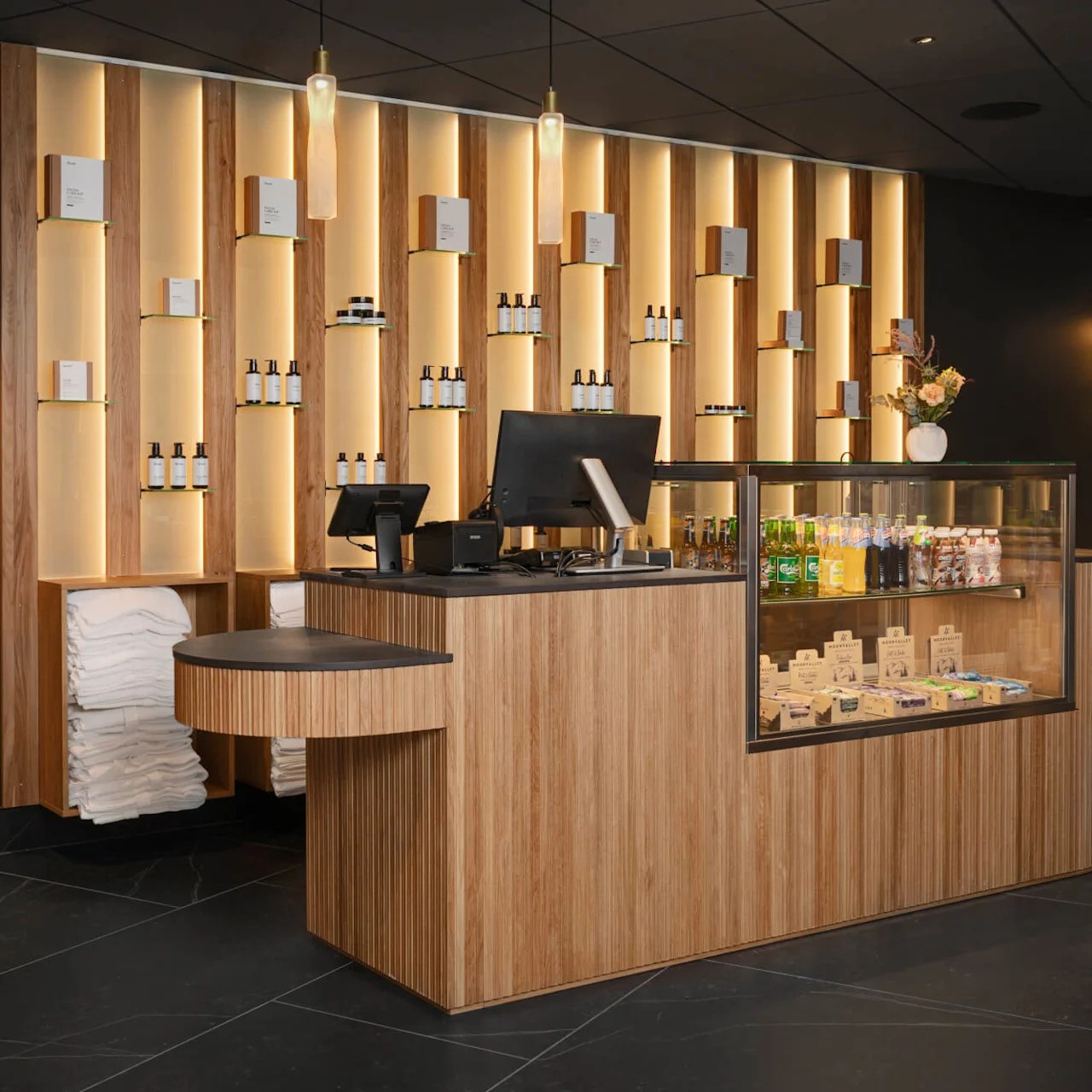 En trädisk i receptionen visar en dator, produkter och en glasmonter med drycker och snacks. Väggställen håller toalettartiklar och handdukar. Modern interiör med varm belysning och mörkt golv.