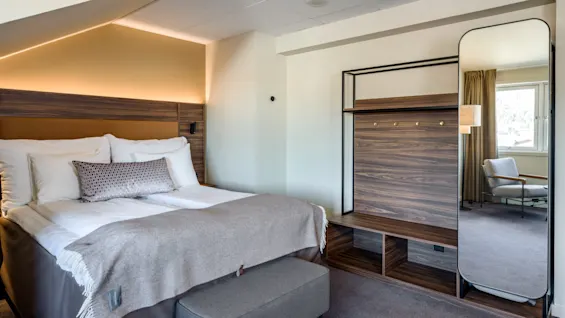 Bed adorned with pillows and a blanket sits in a modern room. A large mirror and wardrobe frame the space. A window admits natural light onto a chair.