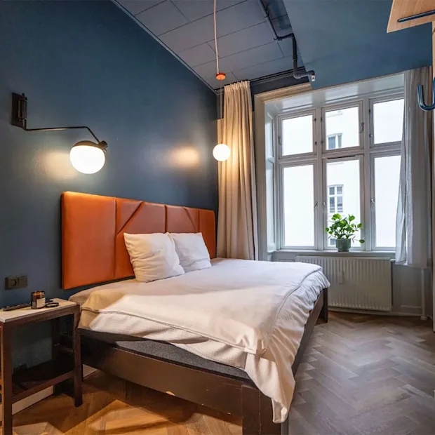 A modern hotel room with a double bed, blue walls, wooden floors, and a large window letting in natural light.