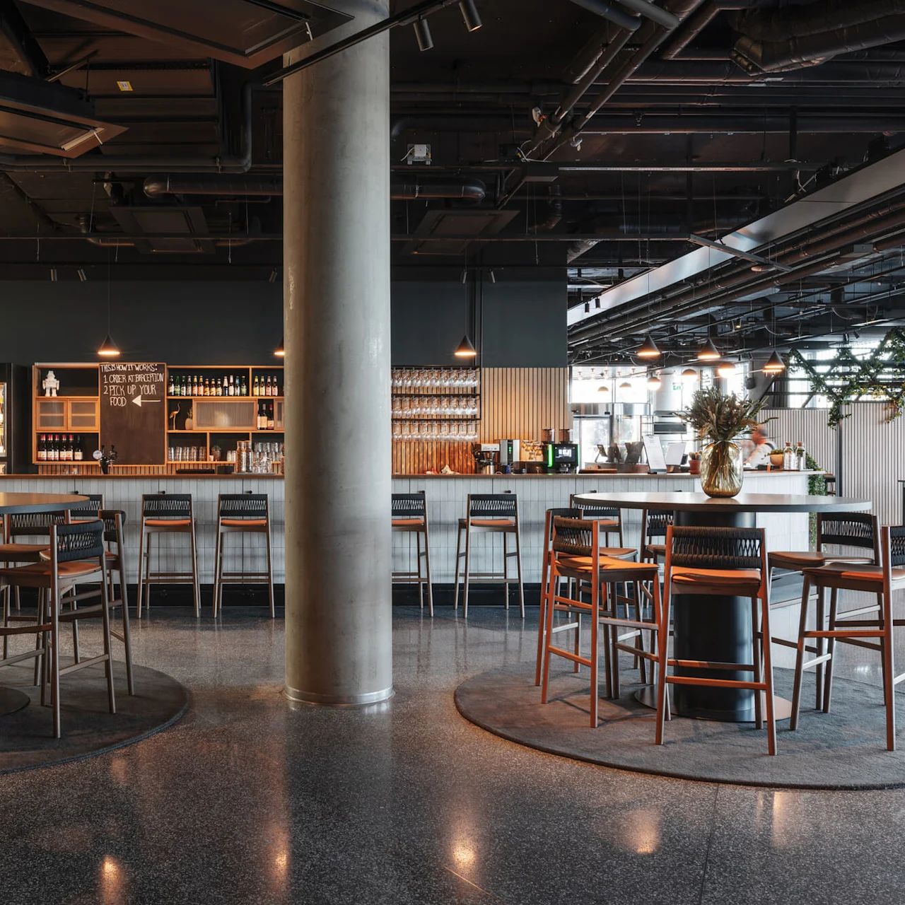 Bar counter with high chairs stands in a modern lounge. Pendant lights hang above, and a chalkboard reads: "1. Order at bartender, 2. Pick up your food here." 