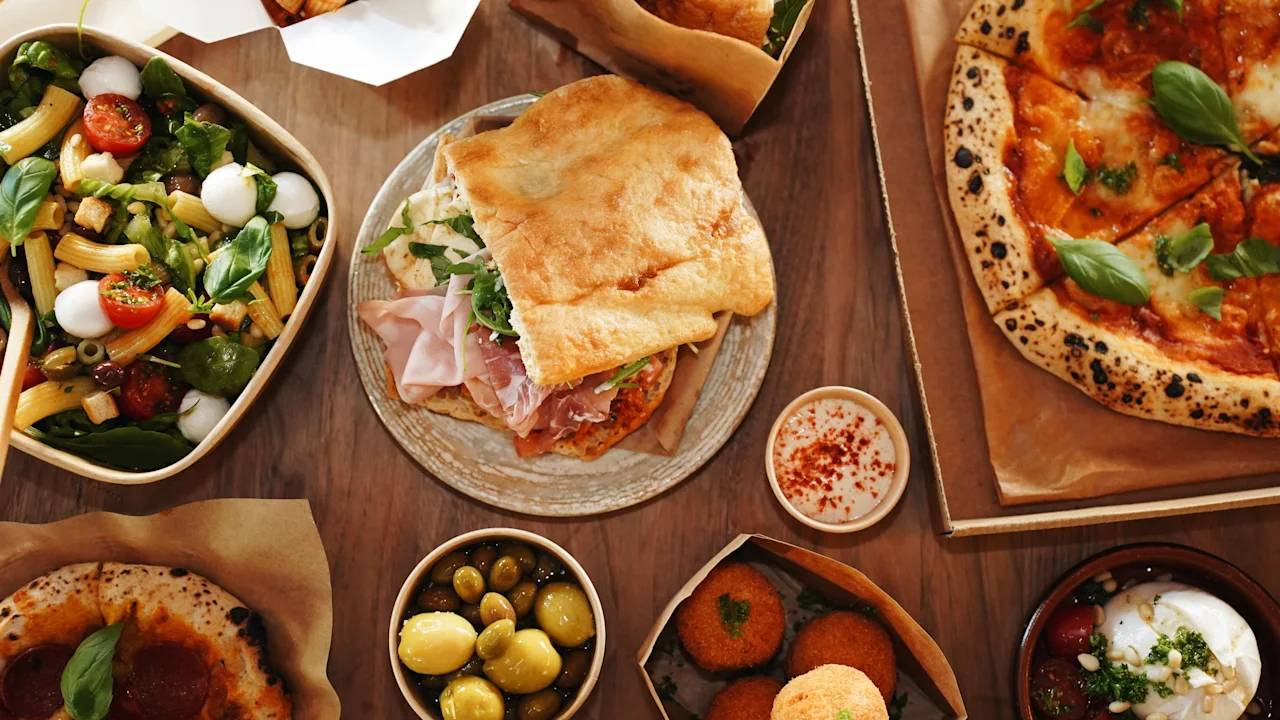 Erilaisia italialaisia ruokia on levitetty puupöydälle, mukaan lukien pizza, voileipä, salaatti, oliivit ja arancini, luoden elinvoimaisen ja houkuttelevan ateria-asetelman, joka on tyypillinen italialaiselle keittiölle.