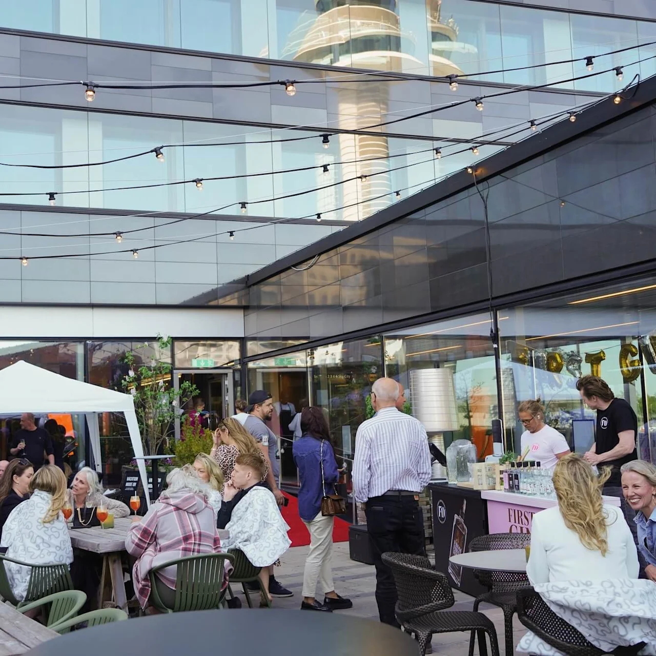 People gather on a rooftop terrace, socializing and drinking at tables. String lights hang above, and a bartender serves drinks under a “FÖRSTA” banner near glass doors. 