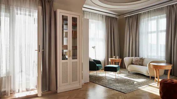 Transparente gardiner slipper lys inn i en koselig stue med beige sofa, grønne stoler, en glassfront bokhylle og runde trebord på et mønstret teppe.