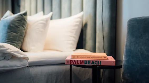 Böckerna med titlarna "The Secret Book of Coastal Living" och "PALM BEACH" ligger på ett litet bord bredvid en säng med fluffiga kuddar och en sammetssänggavel i ett mysigt sovrum.
