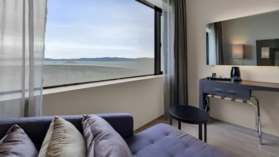 Sofa with pillows facing large window overlooking mountains; adjacent is a wooden desk with a lamp, coffee maker, and tray. Light curtains frame the window. No text present.