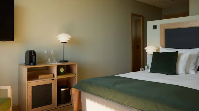 A neatly arranged hotel room features a bed with green accents, a wooden shelf with a lamp and a coffee machine. Neutral walls and warm lighting create a peaceful atmosphere.