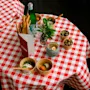 Rödvit-rutigt bordskläde med brödpinnar, flaskvatten, oliver, växt, ljus och två skålar med mat; omgiven av meny och glas på trägolv. Text på meny: "klassig".