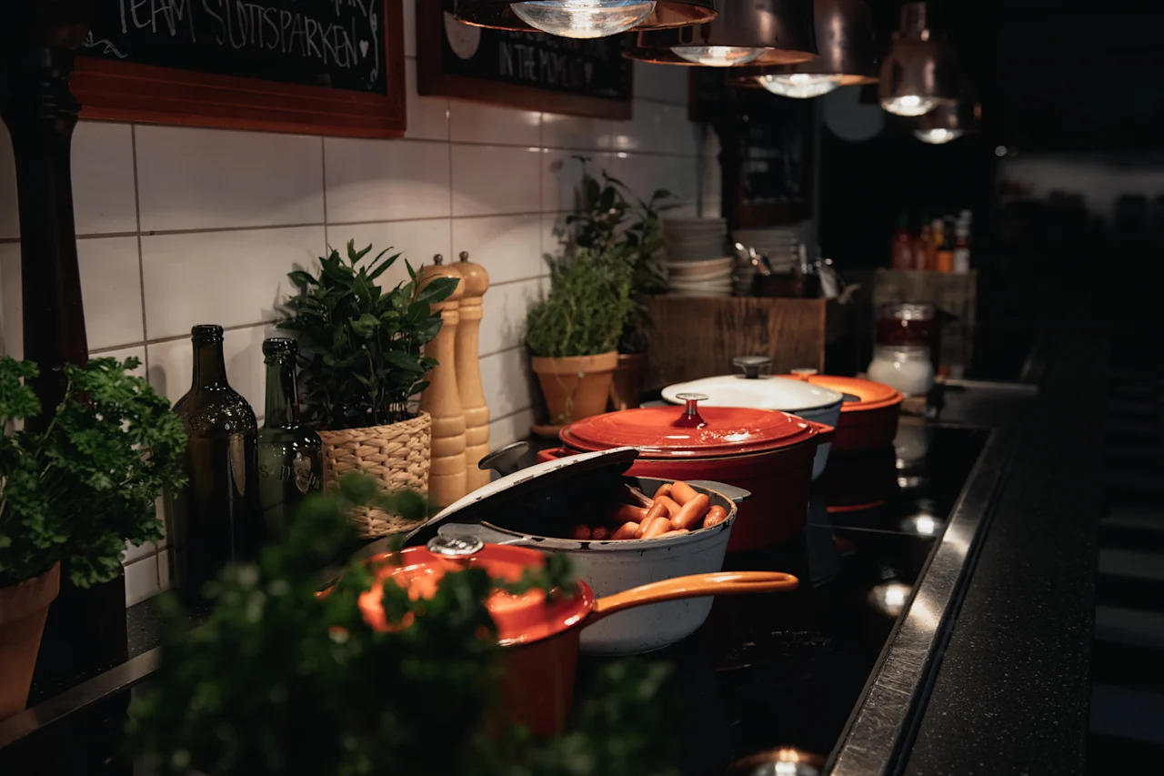 A kitchen counter with cooking pots, one holding sausages. Green plants and bottles are visible. Signs read TEAM SUITSPARKEN IN THE MOMENT.