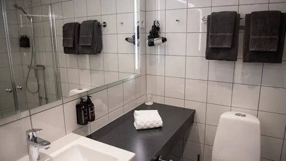 Handfat med kran bredvid vikta handdukar på en mörk bänkskiva i ett vitkaklat badrum; handdukar och toalettartiklar är prydligt organiserade, och en glasdusch är synlig.