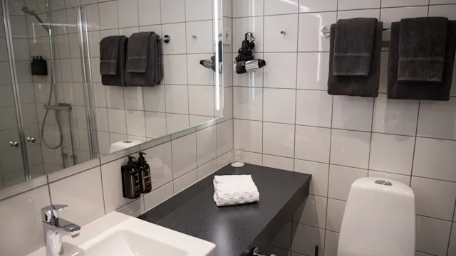 Sink with faucet sitting beside folded towels on a dark countertop in a white-tiled bathroom; towels and toiletries are neatly arranged, and a glass shower is visible.