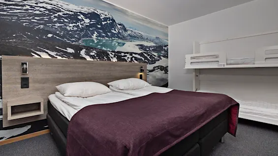 En dubbelsäng med vita kuddar och lila filt, intill våningssängar, i ett rum med en stor väggmålning av berg och sjö.