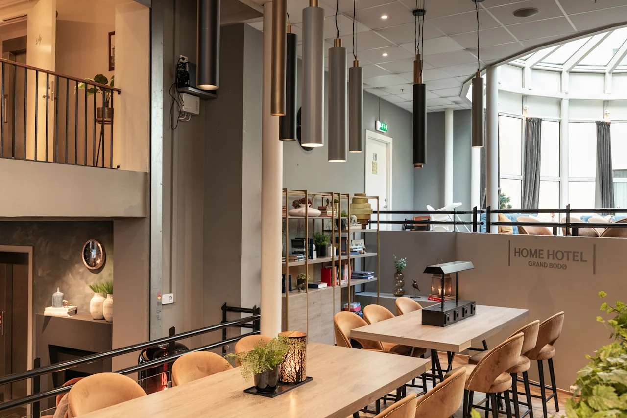 A modern and inviting multi-level common area at Home Hotel Grand Bodø with dining tables, stylish lighting, and a bright window.