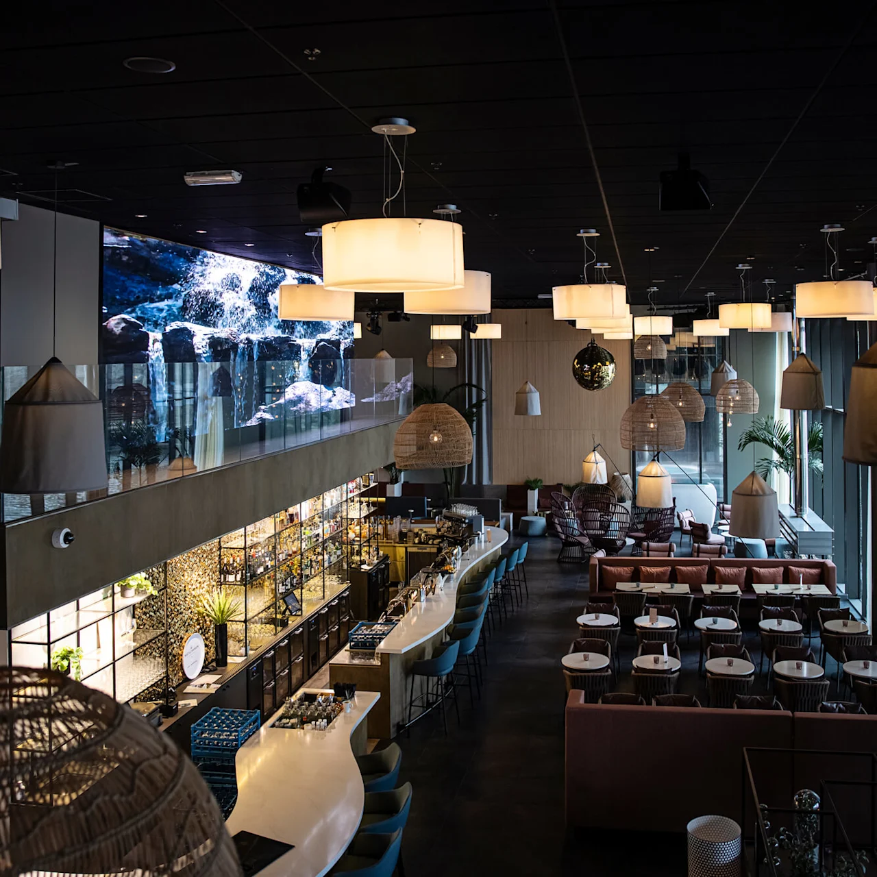 En stilfull restaurantinteriør med moderne sitteplasser, hengende pendellamper og en lang, buet bar. Store vinduer og veggskjermer som viser fosser skaper en elegant stemning.