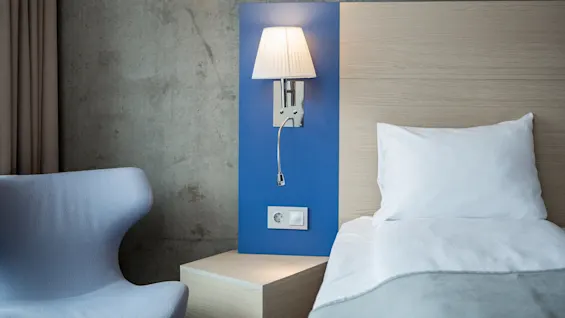 A wall-mounted lamp illuminates a neatly made bed with white linens. A light blue chair and a wooden nightstand are nearby, creating a modern and minimalist bedroom setting.