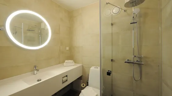 A modern bathroom features a lit circular mirror above a long white countertop with a faucet. Adjacent, a toilet is placed. A glass-enclosed shower is in the corner.
