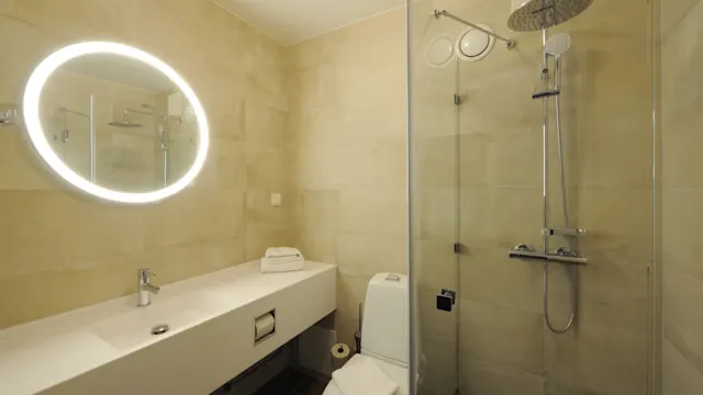 A modern bathroom features a lit circular mirror above a long white countertop with a faucet. Adjacent, a toilet is placed. A glass-enclosed shower is in the corner.