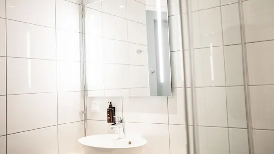 Handfat med en tvålpump finns under en väggmonterad spegel, omgiven av vita kakelväggar i ett badrum. Ingen text finns.