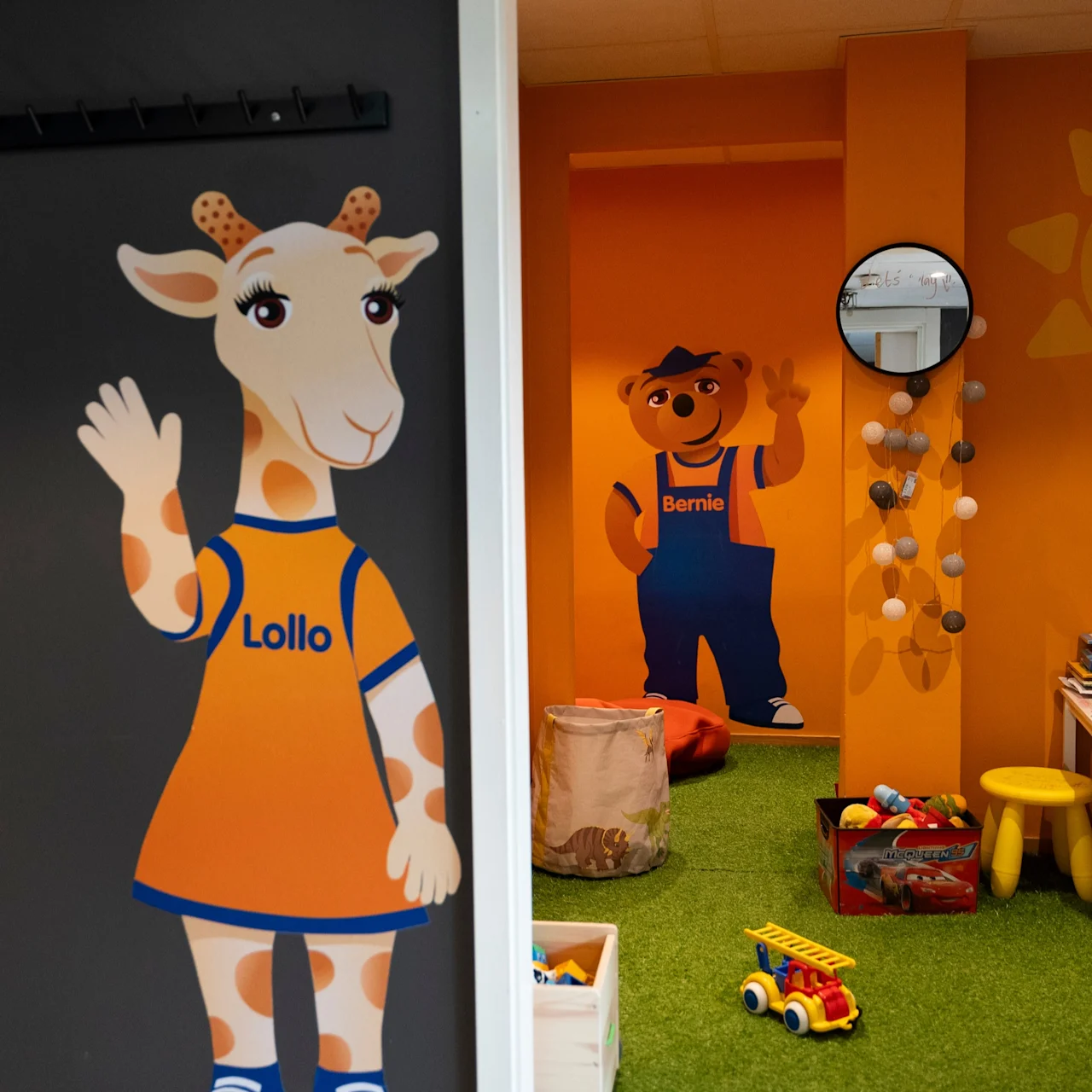 Tecknad giraff och björn på målade väggar. Giraffen, märkt "Lollo," vinkar vid en dörr. Björnen, märkt "Bernie," står i ett färgglatt lekrum med leksaker.