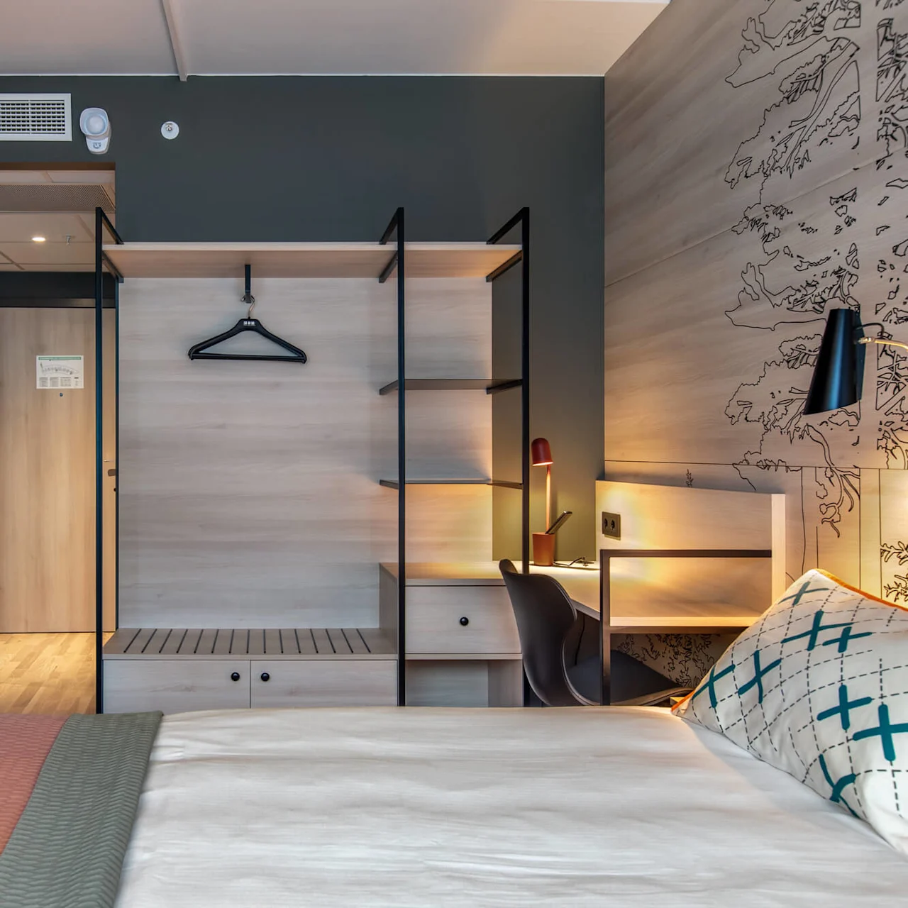 A modern hotel room with a neatly made bed, a wall-mounted shelving unit, and a work desk featuring a lamp. Three colorful hula hoops adorn the wall beside a door.