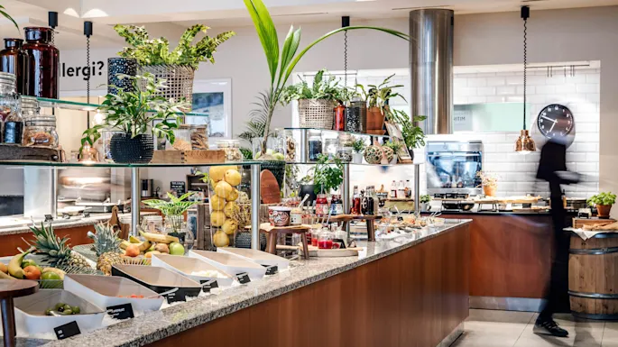 A buffet counter displays various fruits and beverages, accompanied by decorative plants. A blurred person moves past in a modern kitchen setting. Text on the wall: “Allergi?”