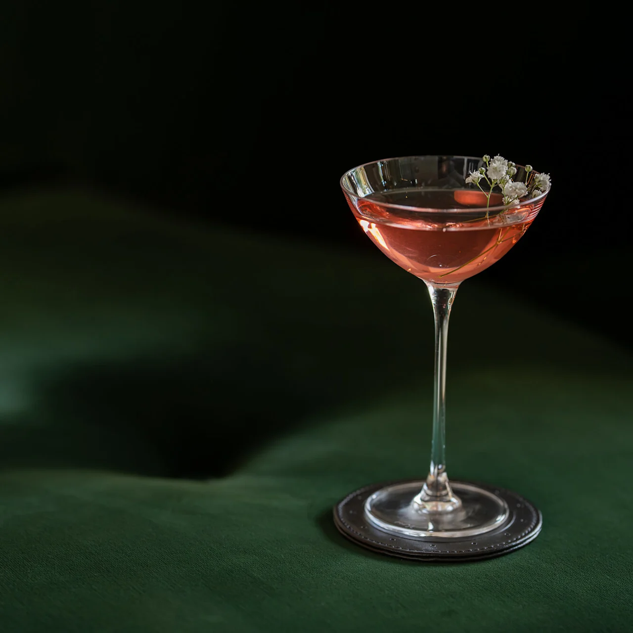 A cocktail glass with pink liquid and white flowers floats on a dark green surface in a dimly lit environment.