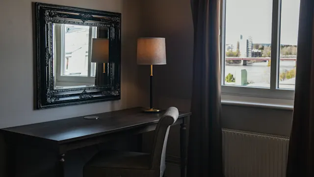 A dark wooden desk holds a lamp and chair, placed by a window with city view. A decorative mirror hangs on the wall, reflecting the lamp.