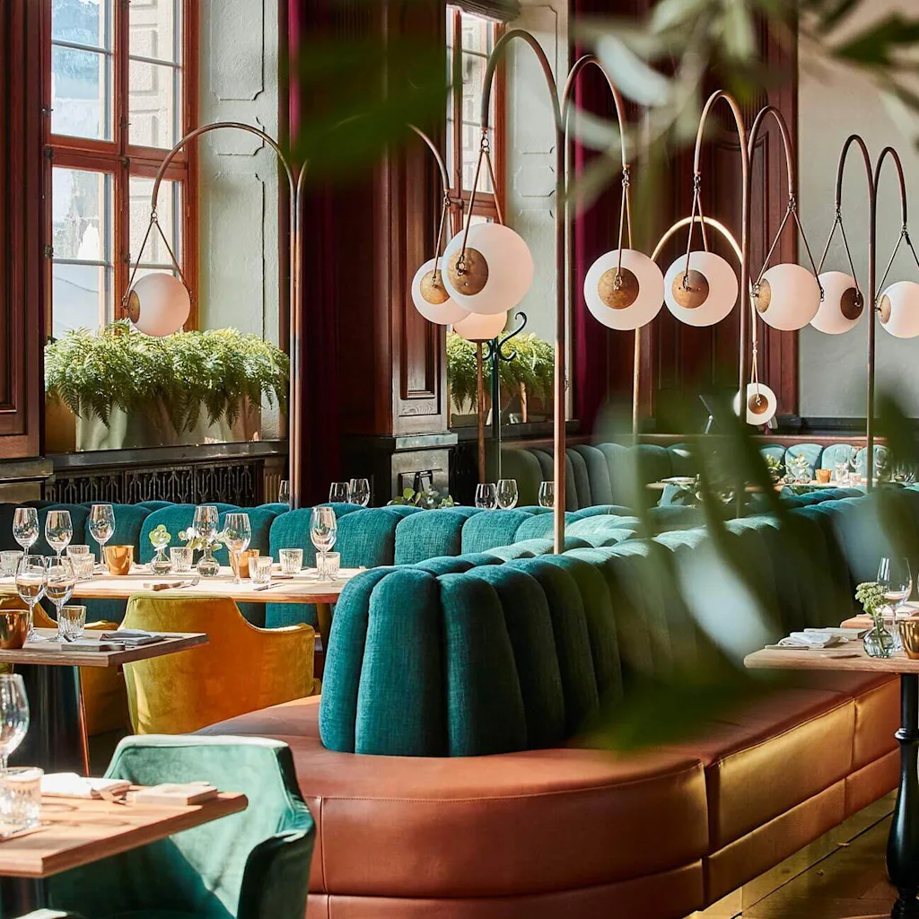 Elegant dining room with plush seating, set tables, hanging lamps, and vibrant plants. Vintage and contemporary elements in a bright, sophisticated setting.