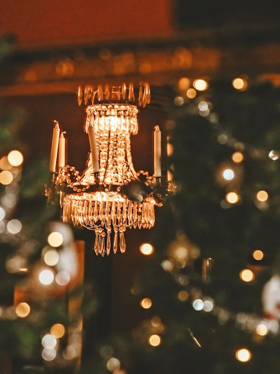 A glowing crystal chandelier hanging in a cozy, festive room decorated with warm Christmas lights.