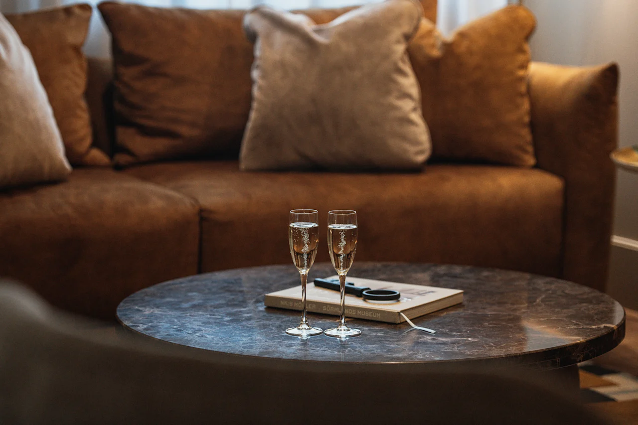Två champagneglas på ett soffbord framför en bekväm soffa, med en bok med titeln KUNST SÖNNDAG MUSEUM.