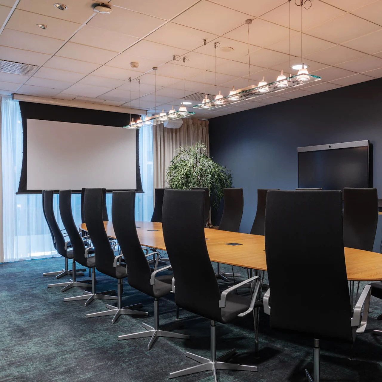 Ett konferensrum har ett långt träbord omgiven av högryggade svarta stolar. En projektionsskärm är framför, med en stor monitor på väggen och en växt i närheten.
