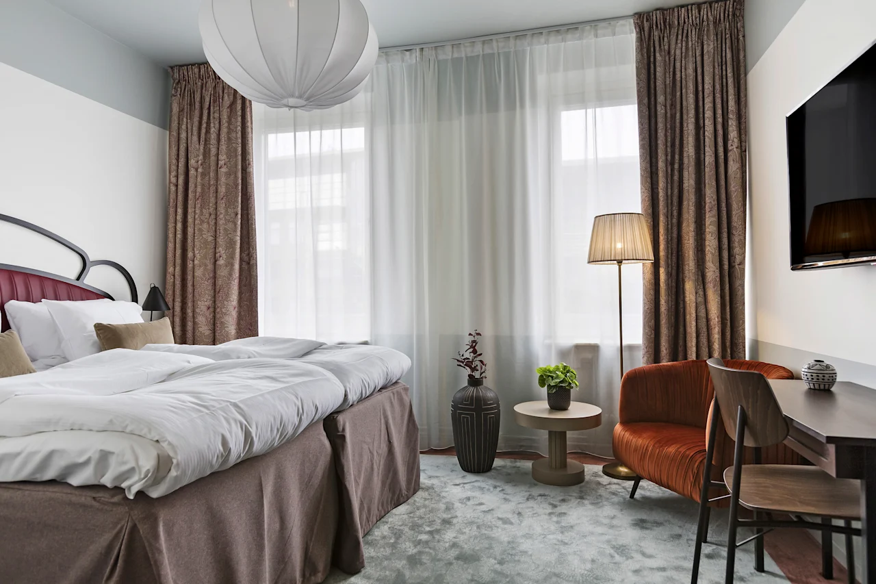 Lyst og koselig hotellrom med oppredd dobbeltseng, sittegruppe ved vinduet og et lite skrivebord.