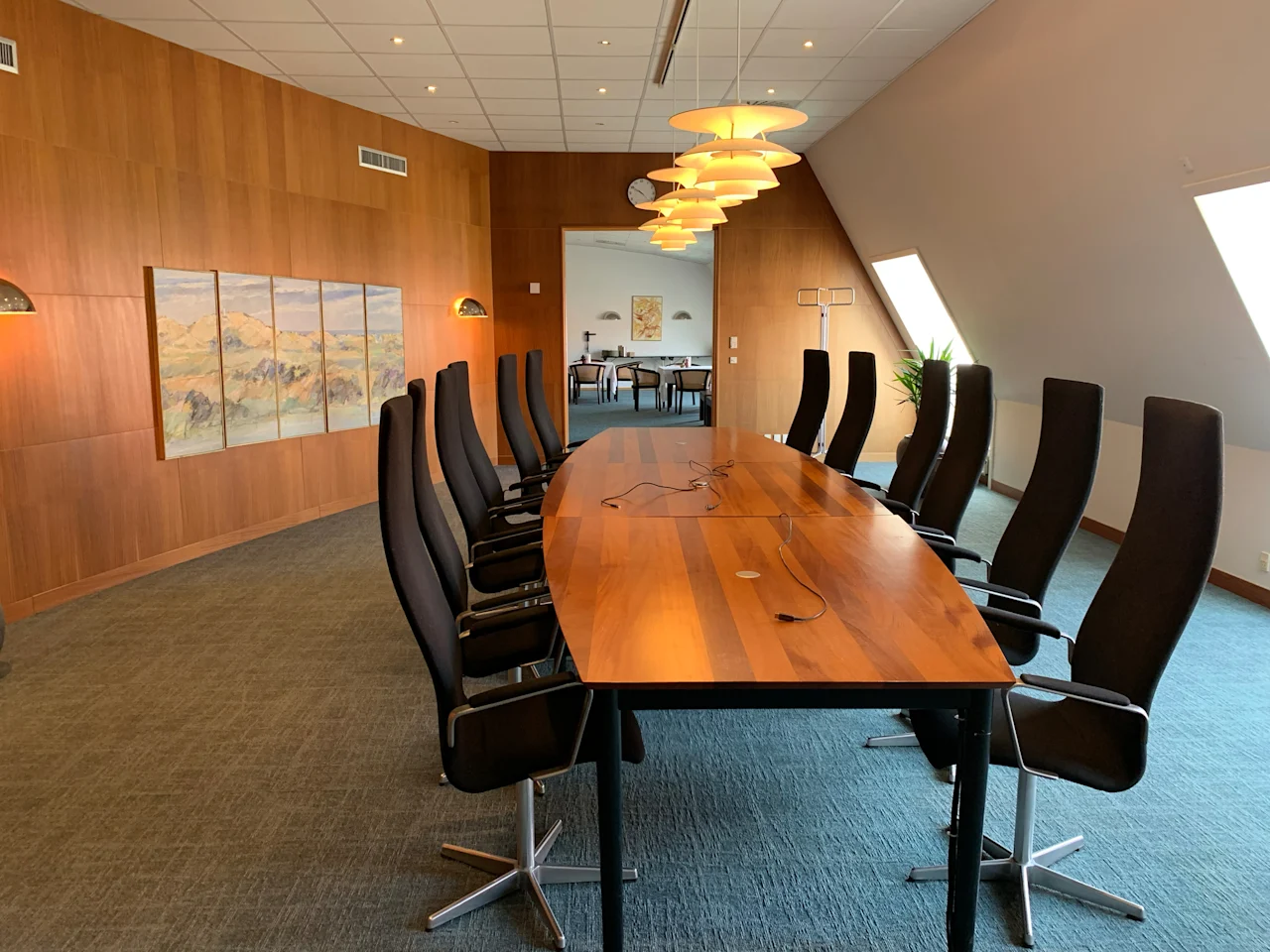 Long wooden conference table surrounded by black chairs, under hanging lamps, in a wood-paneled room with a painting and natural light from slanted windows.