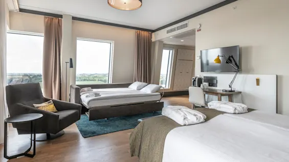 A modern hotel room features two neatly made beds with folded bathrobes. A desk with a chair and lamp is beside a television. Large windows offer a scenic view outside.