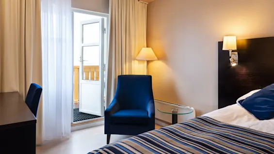 A blue armchair sits beside a floor lamp in a cozy room. A bed with striped bedding is nearby. An open door leads to a balcony, surrounded by light curtains.