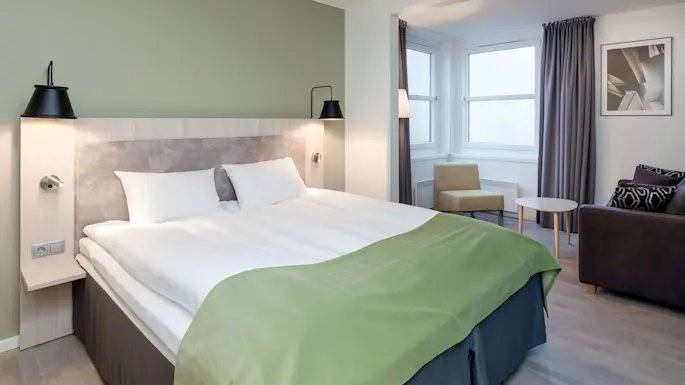 Bed with white sheets and green throw sits against light green wall, flanked by black lamps. A chair and table populate the room's corner by curtained windows.