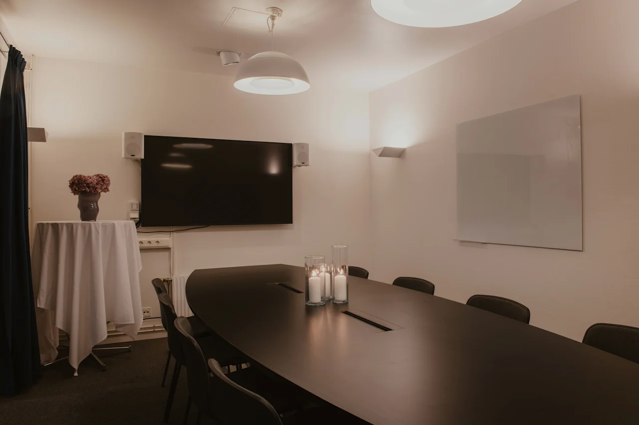 Et veloplyst konferencelokale med en stor skærm, et langt mødebord og en whiteboard, klar til præsentationer eller diskussioner.