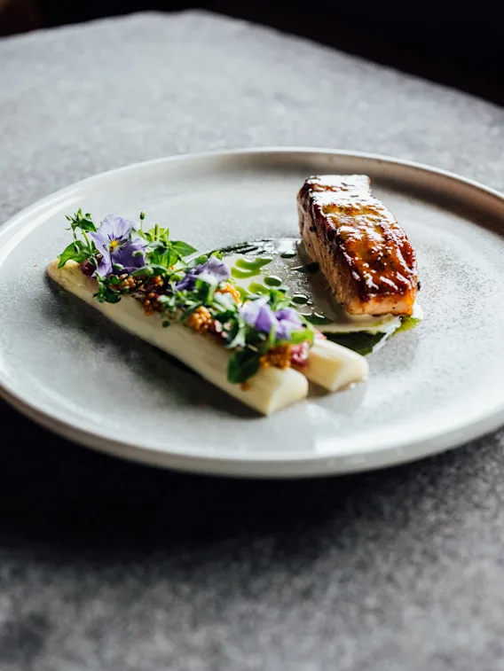 En gourmetskål med stekt fisk ved siden av hvit asparges pyntet med urter og lilla blomster, på en strukturert grå tallerken i dempet belysning.
