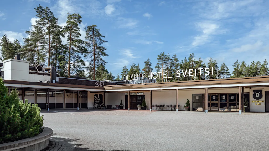 Suuri hotelli, jossa lukee "Quality Hotel Sveitsi" näkyvästi julkisivussa. Ympärillä laaja ajotie ja puita, taustalla kirkas taivas.