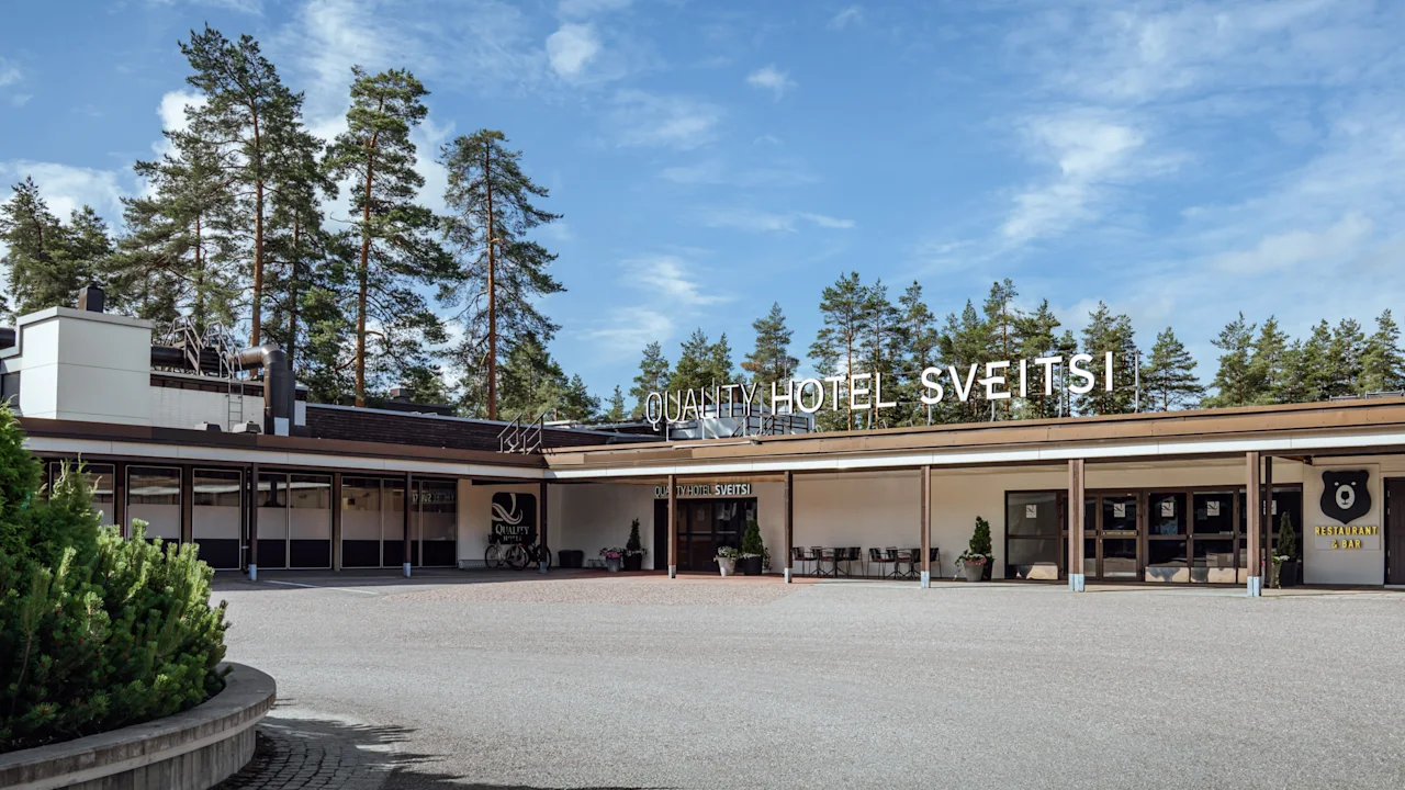 Suuri hotelli, jossa lukee "Quality Hotel Sveitsi" näkyvästi julkisivussa. Ympärillä laaja ajotie ja puita, taustalla kirkas taivas.
