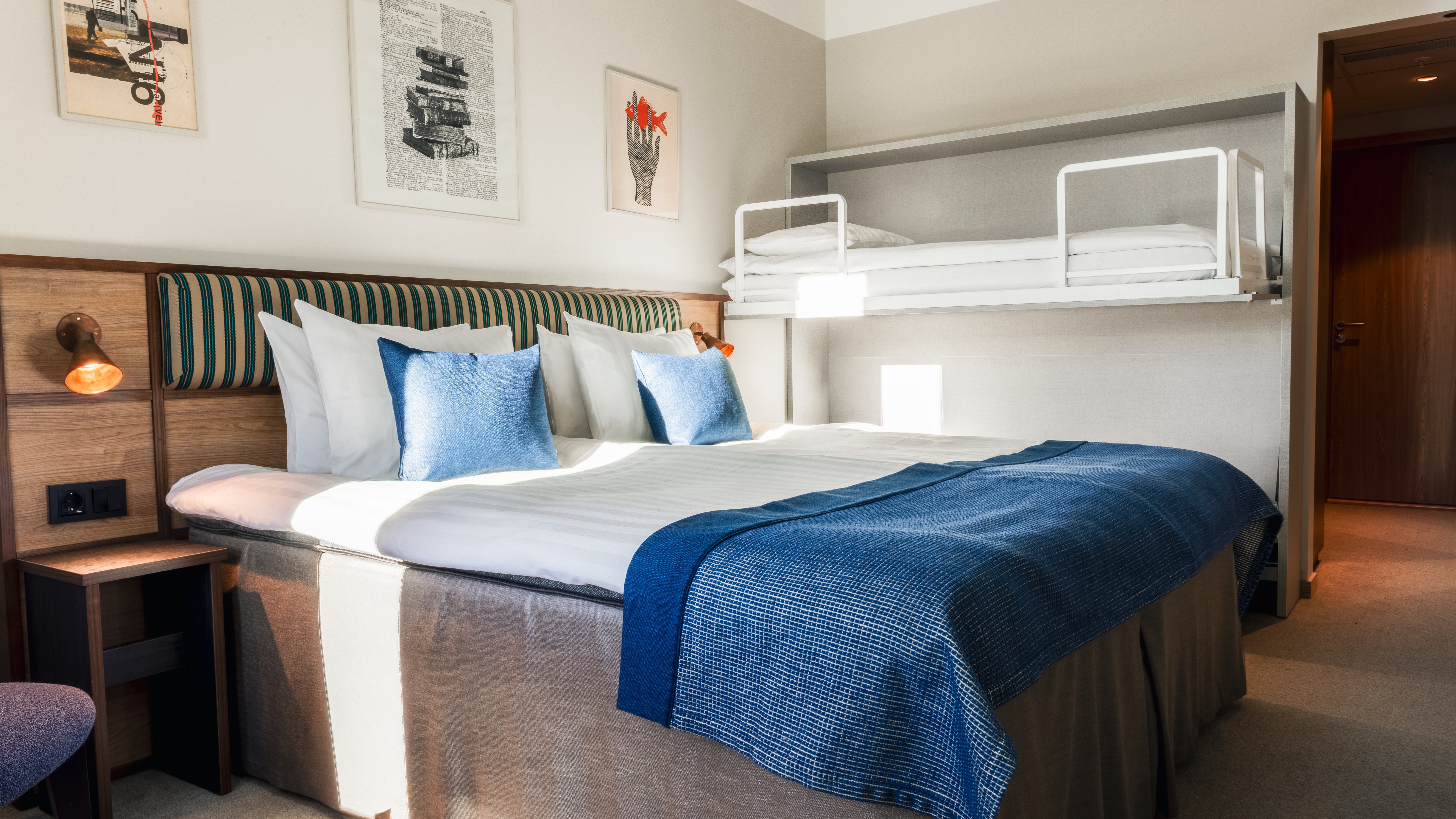 Et hyggeligt hotelværelse med en stor seng og en vægmonteret køjeseng, med blåt og hvidt sengetøj og indrammet kunst på væggen.