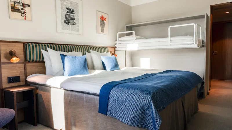 Et hyggeligt hotelværelse med en stor seng og en vægmonteret køjeseng, med blåt og hvidt sengetøj og indrammet kunst på væggen.