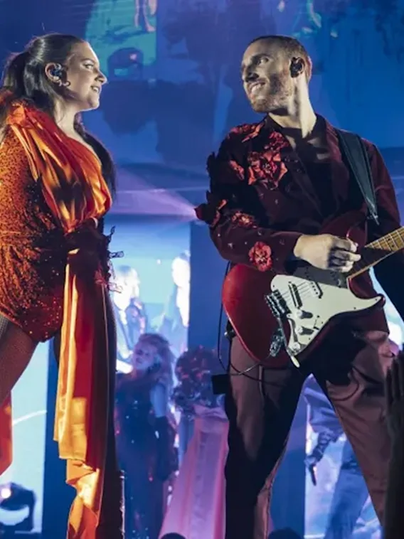 En sanger og en gitarist som opptrer på scenen i fargerike kostymer under fargerikt scenelys.