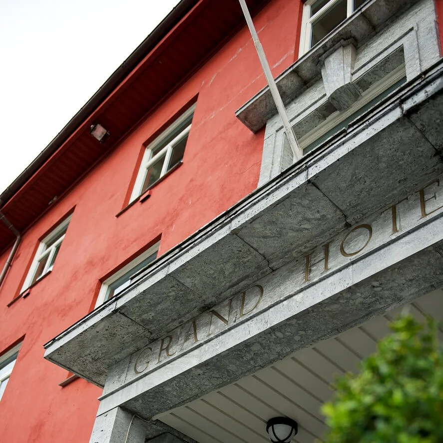 En rød bygning med "GRAND HOTEL" på en marmorinngang, akkompagnert av en gatelykt. Delvis synlig grøntområder er i forgrunnen.