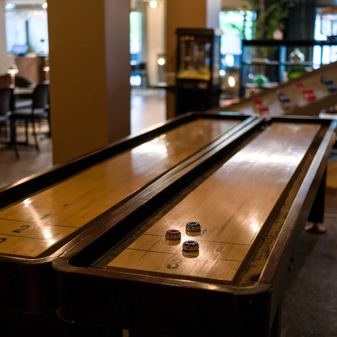 A shuffleboard table with four pucks rests in a dimly lit room surrounded by chairs and various arcade machines.
