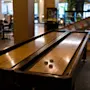 A shuffleboard table with four pucks rests in a dimly lit room surrounded by chairs and various arcade machines.