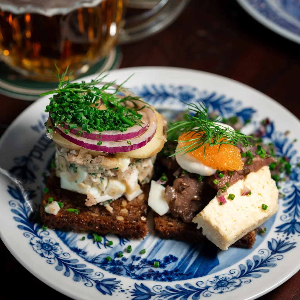 Two delicious open-faced sandwiches with various toppings and a glass of beer on a patterned plate.