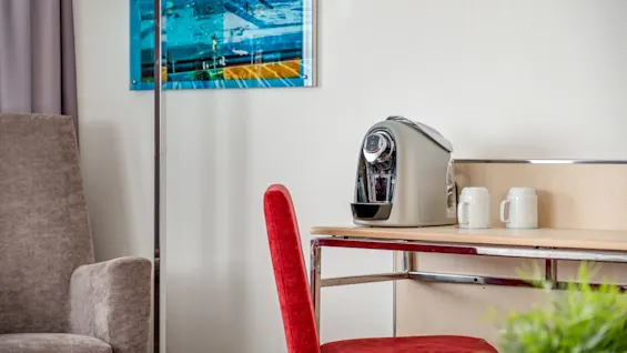 Coffee maker on a table next to two mugs with a red chair, in a room with wall art.